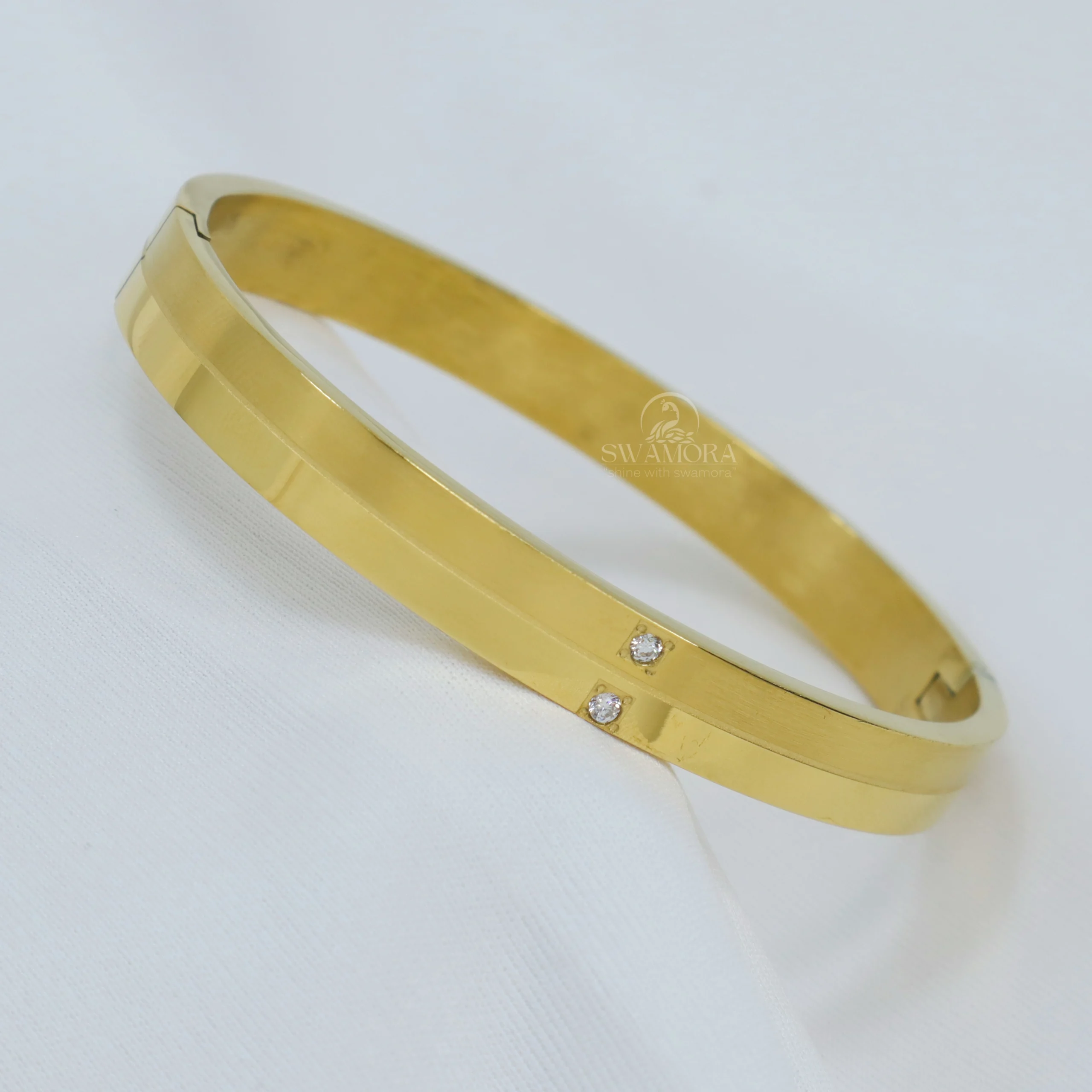 Introducing a chic and modern gold-plated hand kada! Its round design and the two small sparkling stones set on it give it a unique and stylish look. This kada is made from high-quality material, ensuring it lasts for a long time. Key Features: Anti-tarnish: The high-quality gold plating prevents it from tarnishing quickly and helps maintain its shine. Skin-friendly: This material is safe for your skin. Long-lasting: Made with good quality materials, this kada is designed to last. Its round design and the sparkling stones on either side give it a modern and elegant appeal. Suitable for both men and women to use as a style statement. A great option for any special occasion or for everyday wear. Add a special sparkle to your personality by wearing this stylish kada!